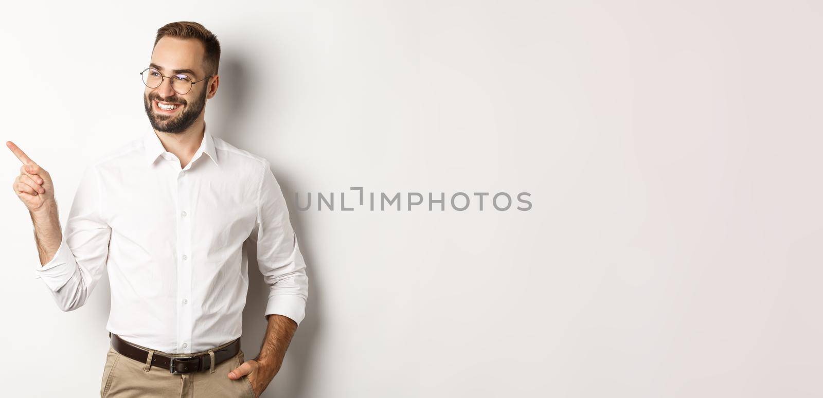 Confident businessman looking satisfied and pointing finger left, showing company banner, standing over white background by Benzoix