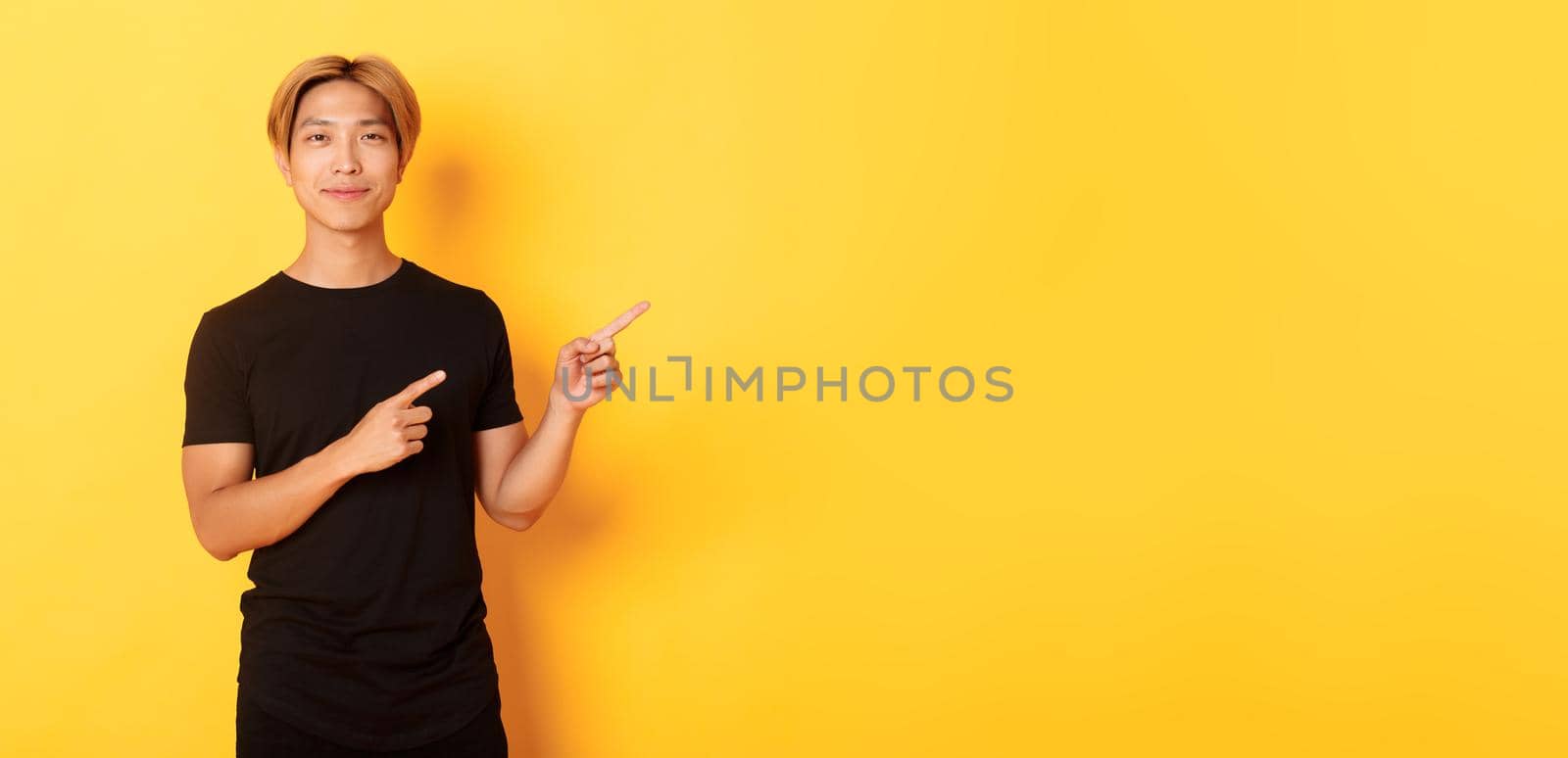 Smiling asian male student with blond hair, pointing fingers right, showing logo, standing yellow background by Benzoix