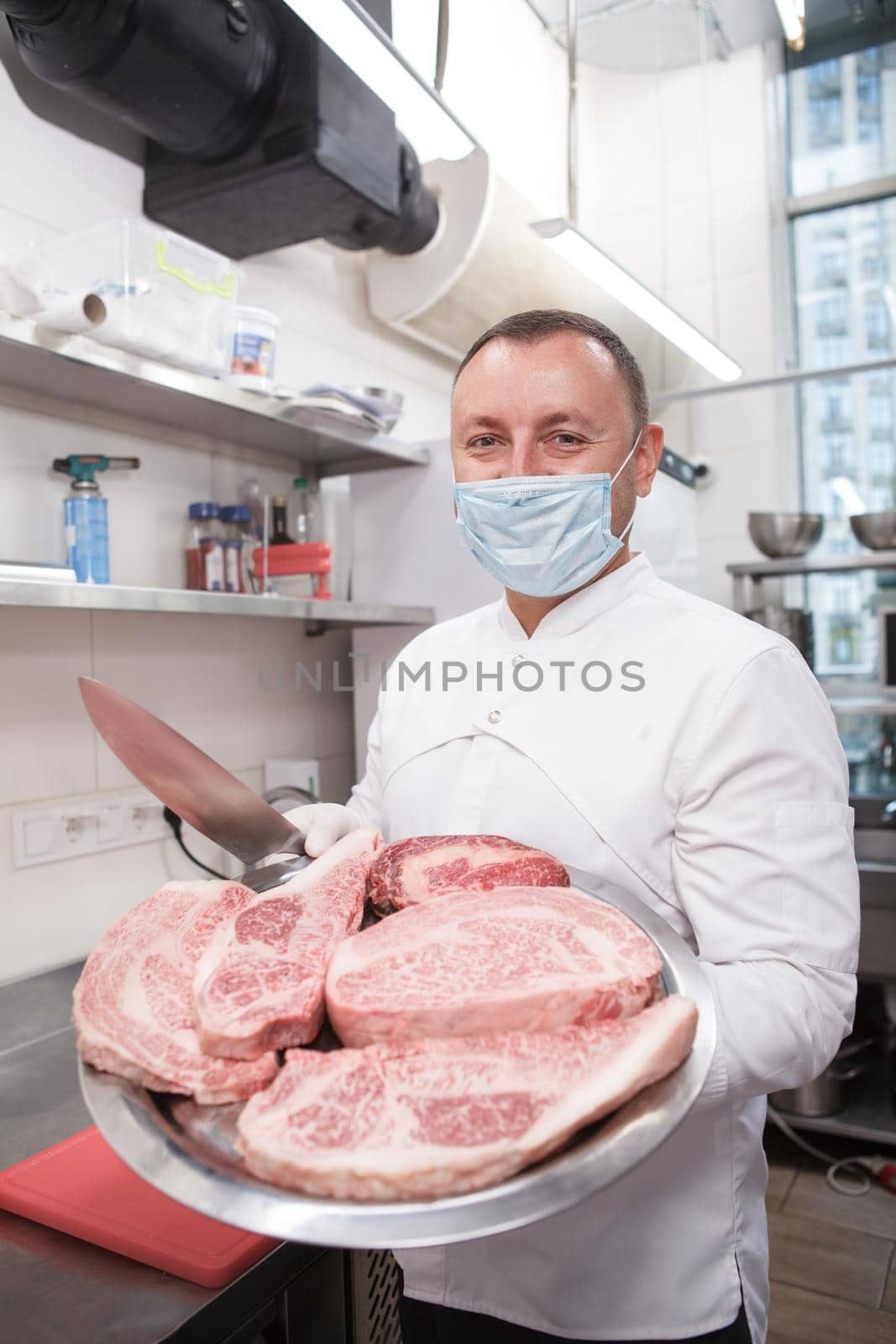 Vertical shot of a professional chef wearing face mask, carrying beef steaks on a tray