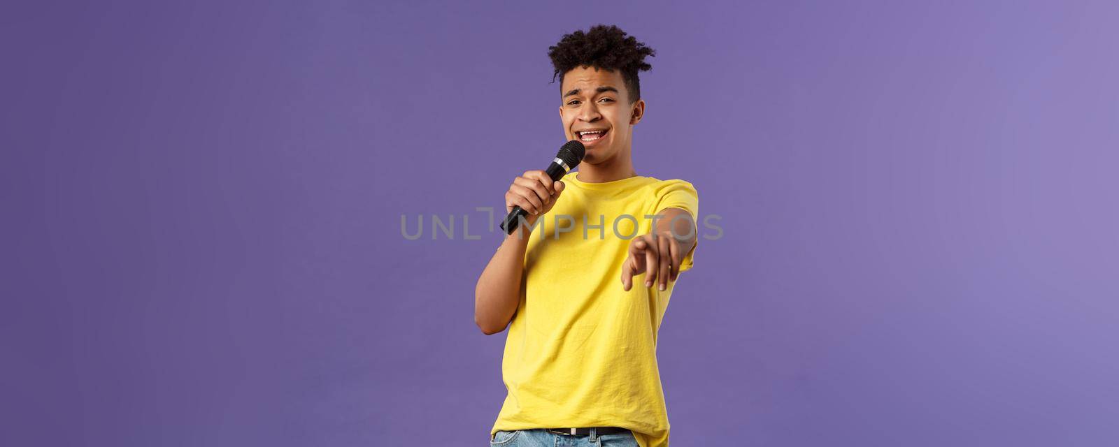 This song is for you. Portrait of romantic carefree hispanic man singing karaoke, pointing at camera as dedicate his performance, holding microphone, standing purple background by Benzoix