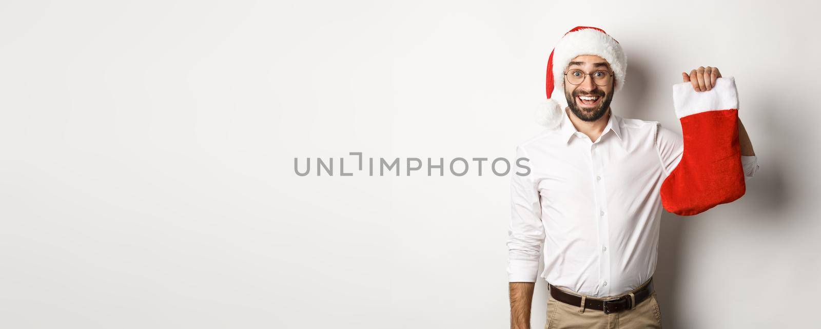 Merry christmas, holidays concept. Excited bearded guy in santa hat holding xmas sock and smiling, celebrating New Year, white background.