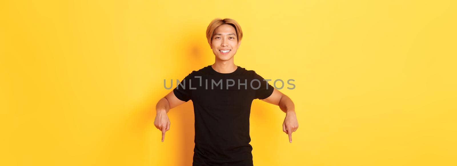 Portrait of smiling attractive korean guy with blond hair, wearing black outfit, pointing fingers down, showing way, standing yellow background.