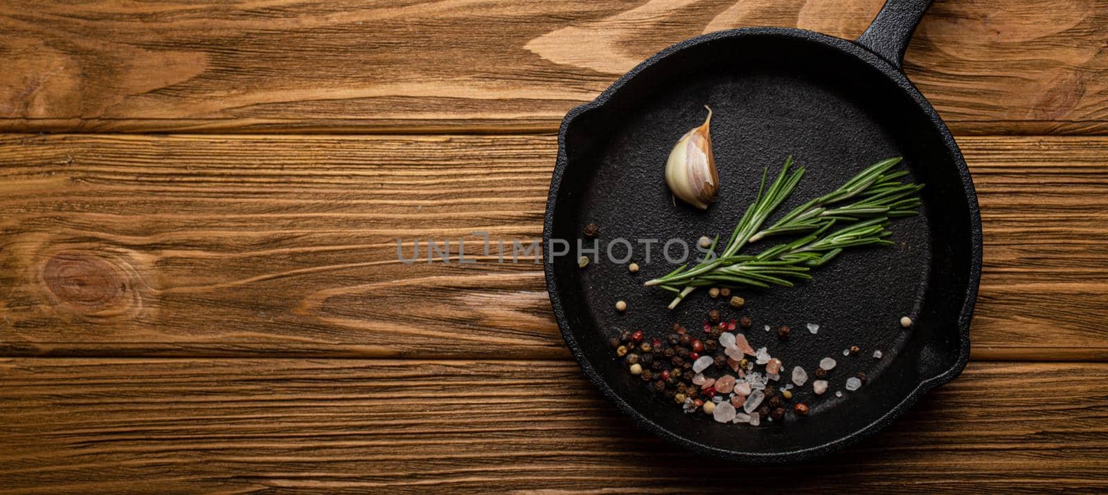 Black cast iron frying pan skillet with food cooking ingredients fresh rosemary, garlic, salt and pepper on rustic wooden table, cooking background and healthy eating kitchen concept copy space