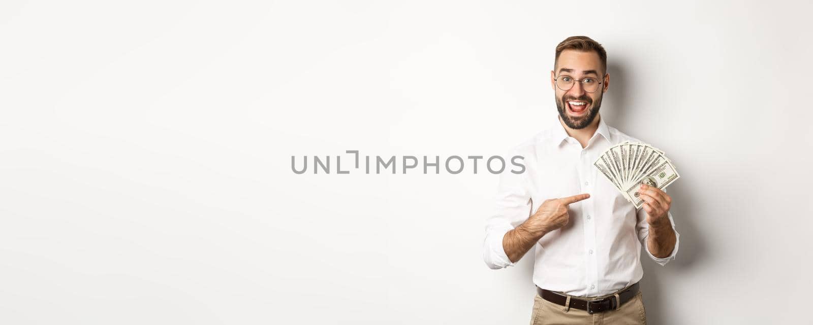 Excited businessman pointing at money, showing dollars and smiling, standing over white background.