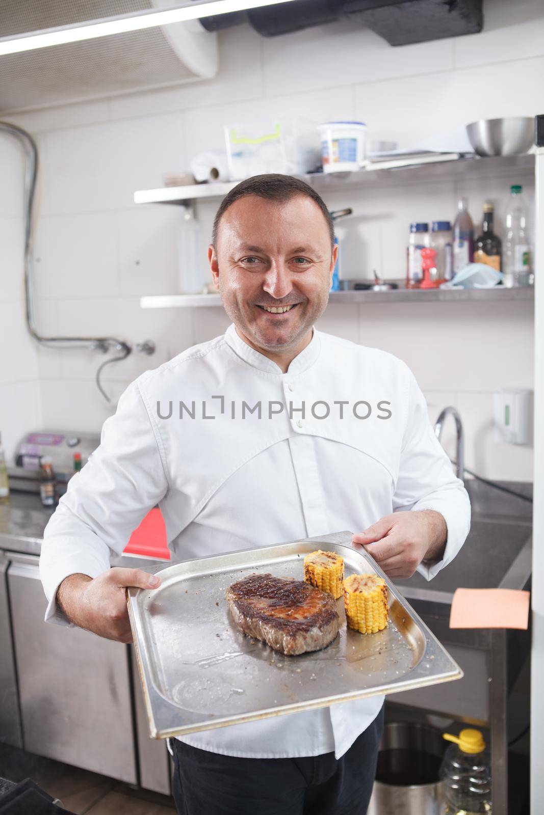 Vertical portrait of a cheerful chef carrying tray with beef steak and sweet corn