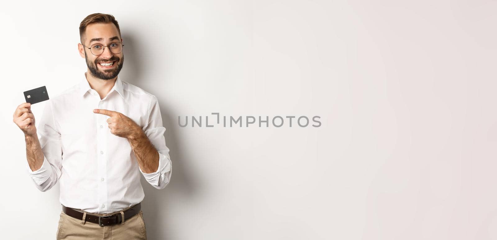 Handsome satisfied man in glasses pointing at credit card, pleased with bank services, standing over white background by Benzoix