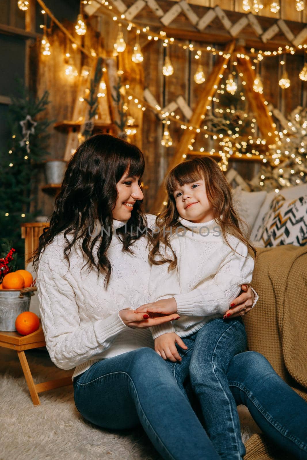 A little girl with her mother in a cozy home environment on the sofa next to the Christmas tree. The theme of New Year holidays and festive interior with garlands and light bulbs.