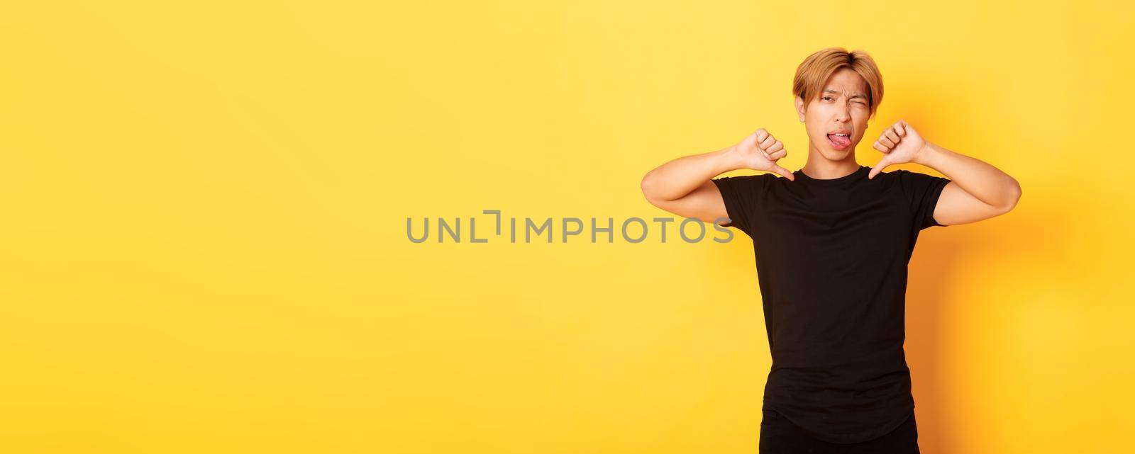 Portrait of sassy asian handsome guy showing thumbs-down and stick tongue disappointed, judge something bad, yellow background by Benzoix