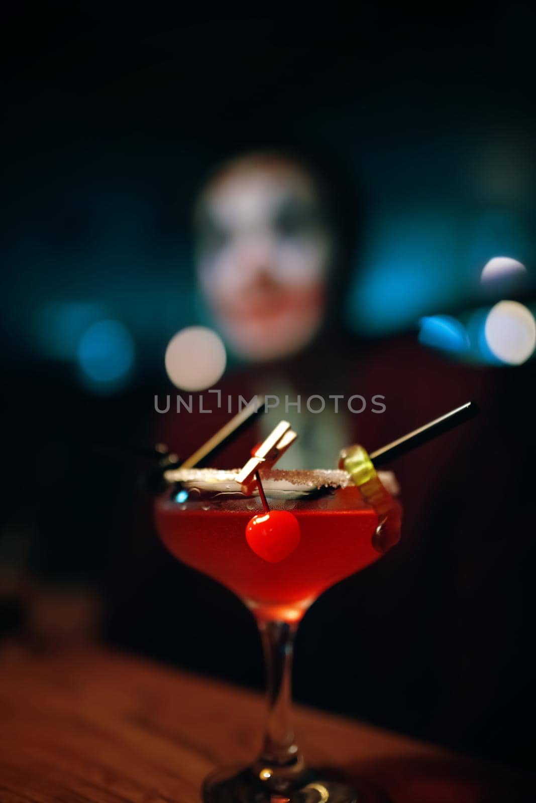 Red cocktail in a glass with a decoration in the form of a marmalade worm and a cherry. Alcoholic drink on the table. Man in clown makeup on the background. Halloween party at a nightclub.
