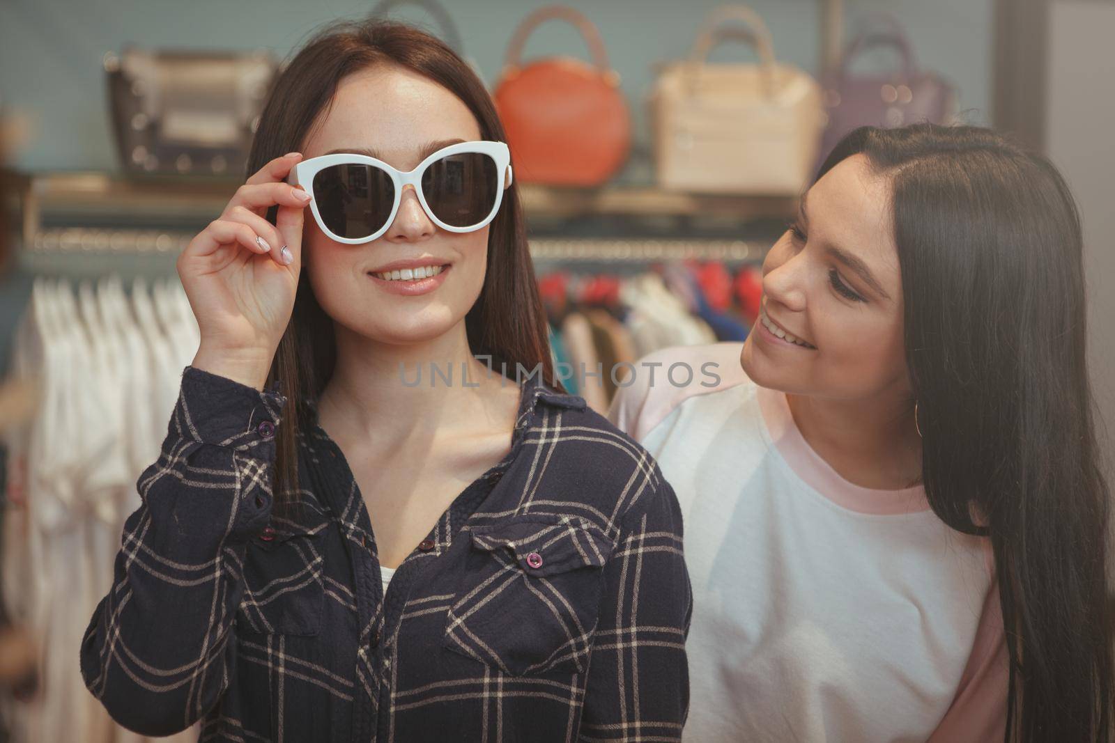 Beautiful woman smiling, trying on new sunglasses, shile shopping with her best friend. Two lovely young women enjoying shopping for eyewear together. Friendship, lifestyle concept
