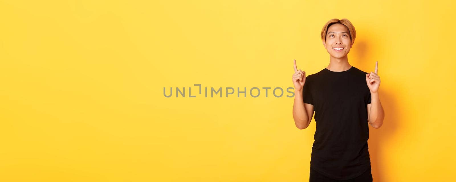 Handsome smiling asian man in black t-shirt pointing fingers up, looking at banner pleased, yellow background.