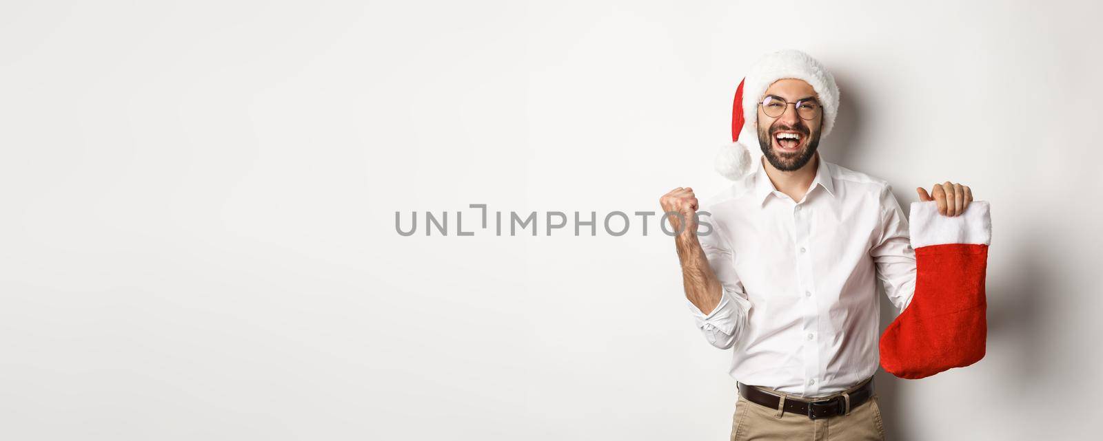 Merry christmas, holidays concept. Happy adult man receive gifts in xmas sock, looking excited, wearing santa hat, white background.
