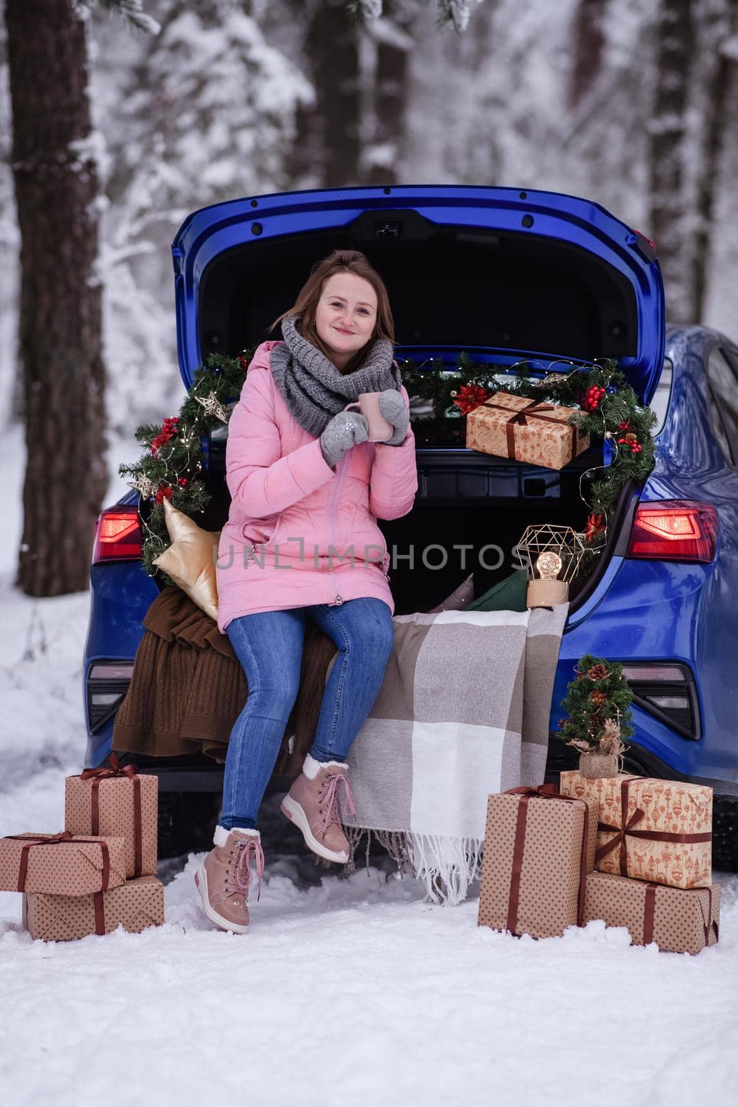 A woman in winter clothes drinks a hot drink sitting in a car decorated in a New Year's way. Traveling by car through the forest. A trip before Christmas.
