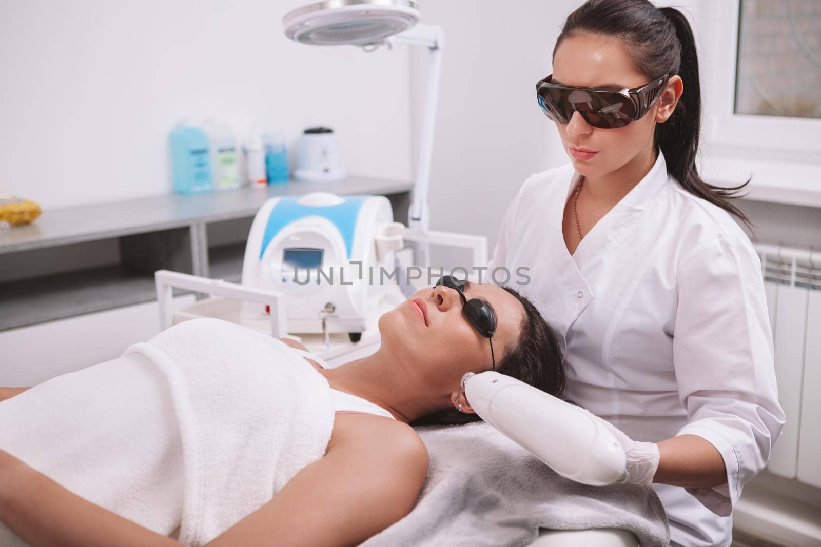 Professional cosmetologist using laser hair removal machine, working with female client. Young woman getting laser skincare treatment at beauty clinic