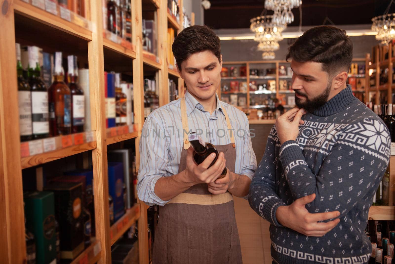 Professional winemaker helping his male customer choosing red wine. Bearded man talking to sommelier at supermarket