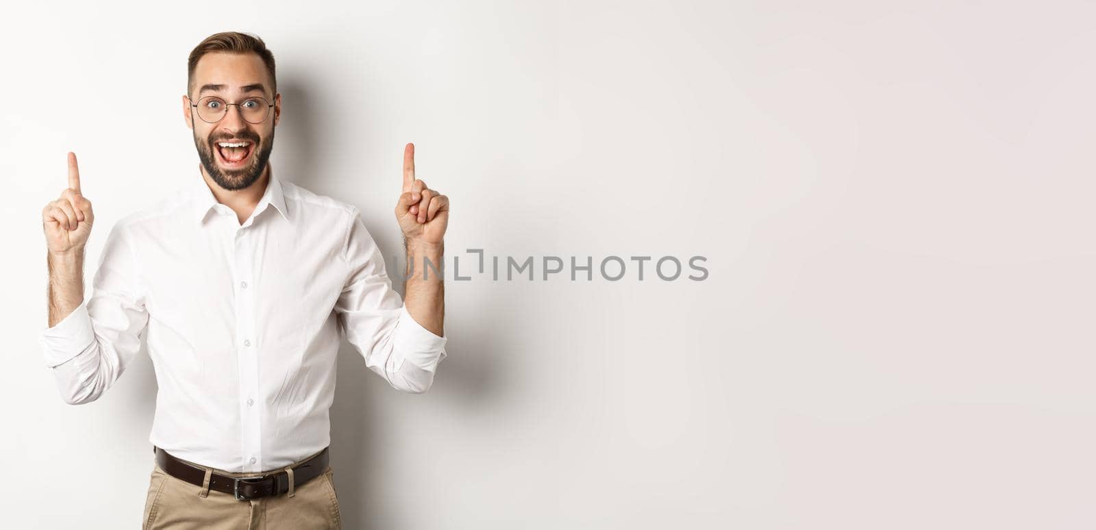 Happy male entrepreneur in business clothes, pointing fingers up and showing promo offer, standing over white background.