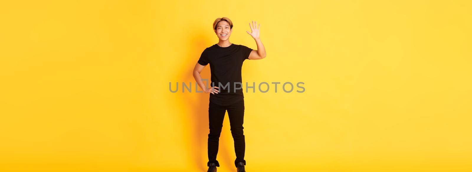 Full-length of friendly handsome asian man waving hand in hello, smiling and saying hi over yellow background by Benzoix