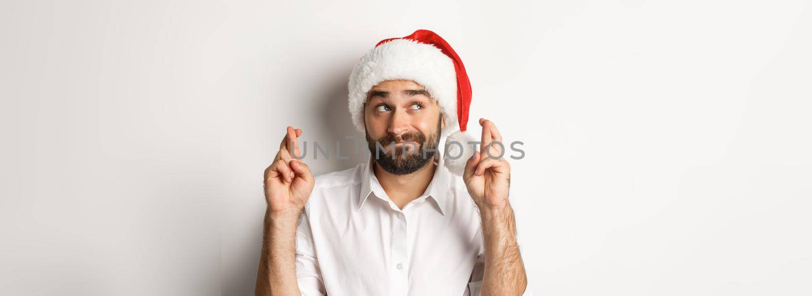 Party, winter holidays and celebration concept. Happy man in santa hat making christmas wish, cross fingers for good luck and looking hopeful at upper left corner.
