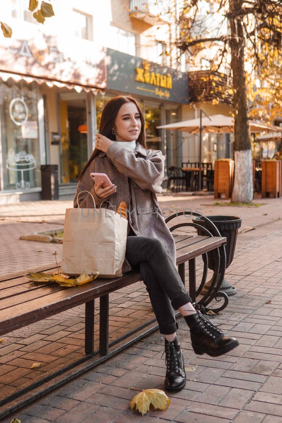 A stylish brunette woman walks around the autumn city. The brunette is sitting on a bench outside with a glass of coffee and a phone, holding a craft bag with a baguette.