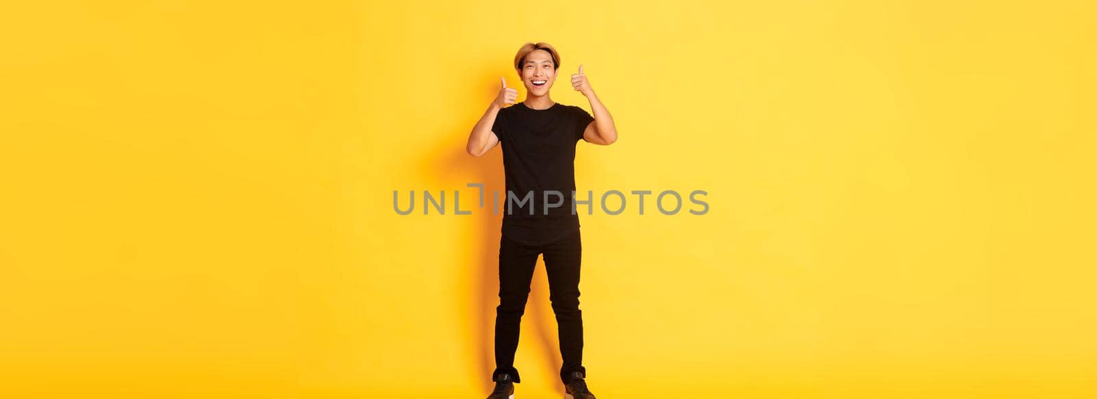 Full-length of satisfied handsome asian guy smiling, showing thumbs-up in approval, yellow background.
