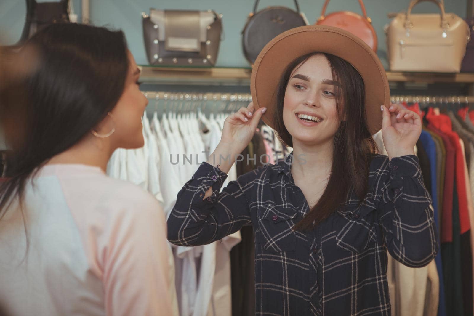 Charming young woman trying on a hat at clothing store, smiling at her friend, asking for opinion. Female friends shopping at clothing boutique together. Consumerism, fashion, sales concept