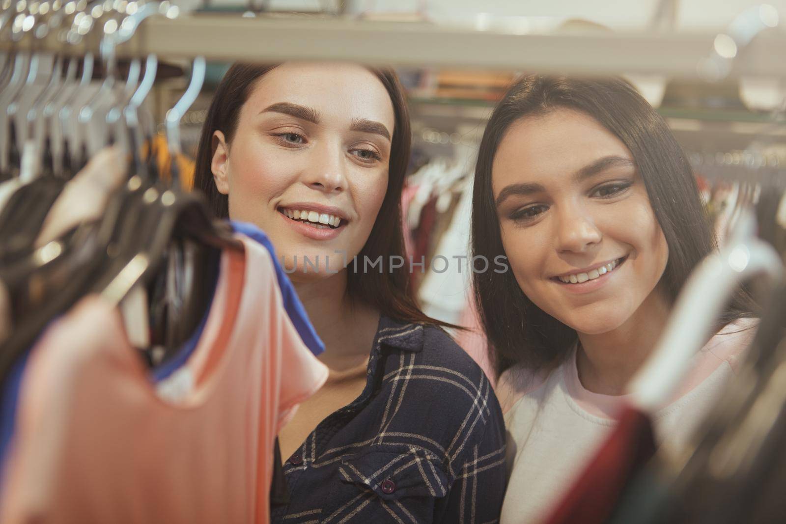 Close up shot of two yougn beautiful women lookign excited, choosing new clothes to buy at fashion boutique. CHeerful female friends enjoying shopping at clothing shop. Retail, buying concept