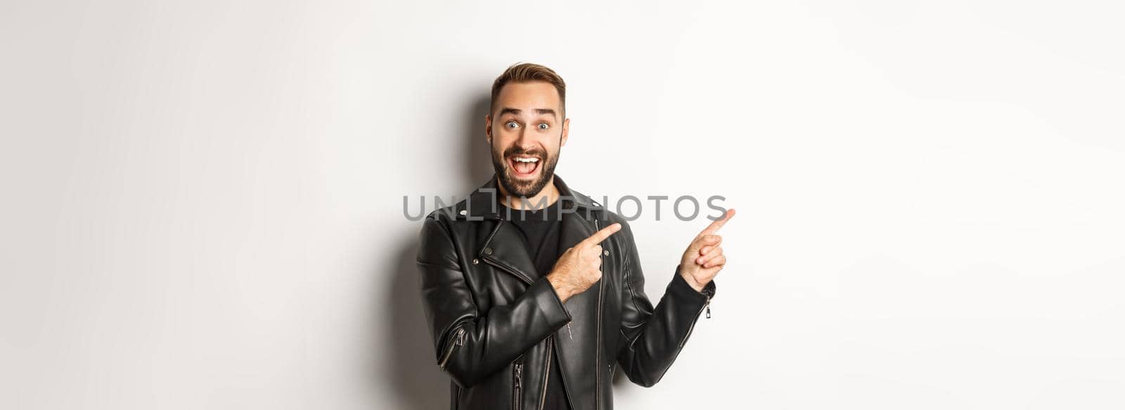 Confident macho man in black leather jacket, pointing fingers left at promo offer, showing logo, white background.