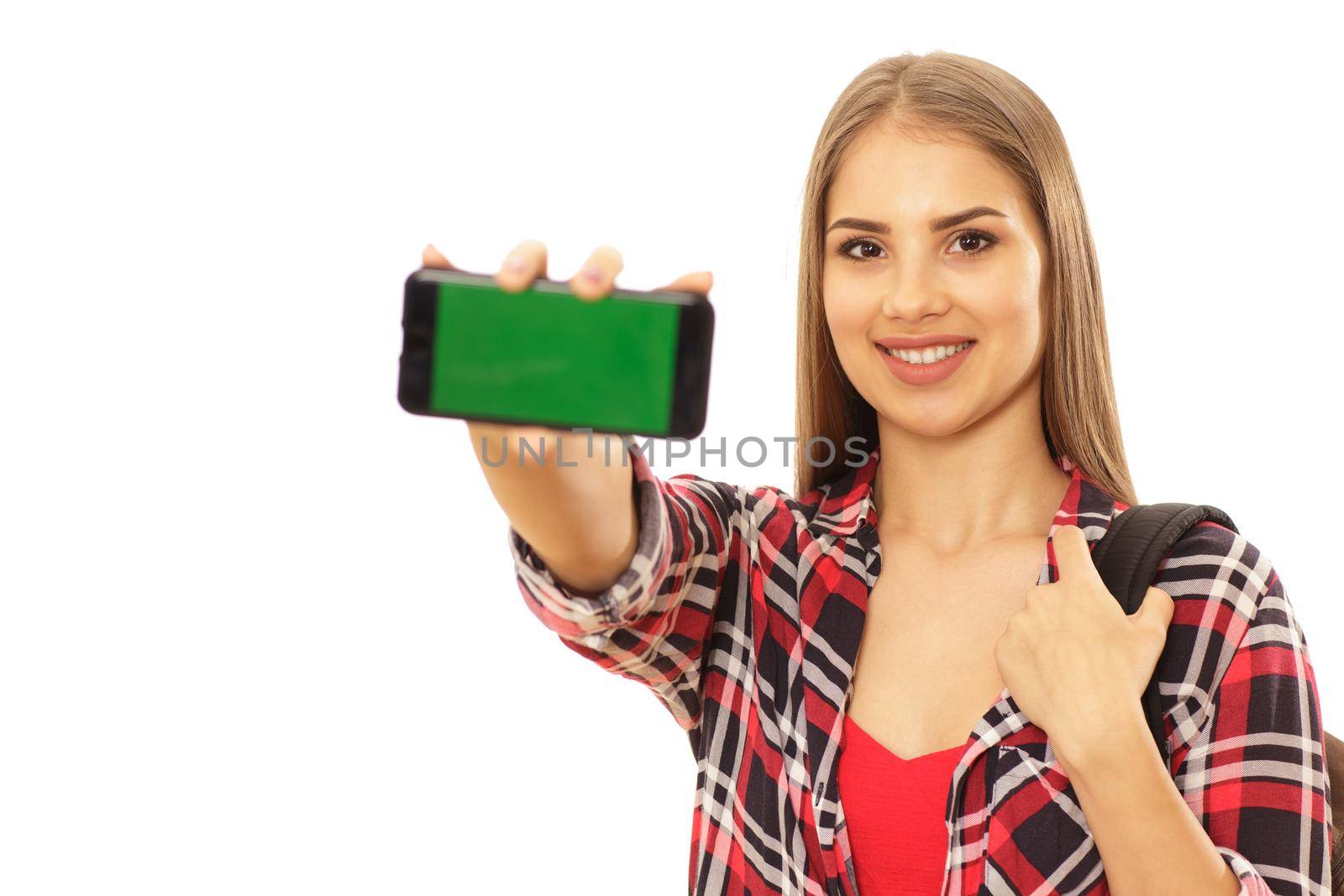 Beautiful female student smiling joyfully, holding out smart phone with copy space green screen to the camera, isolated. Gorgeous woman with backpack holding mobile phone