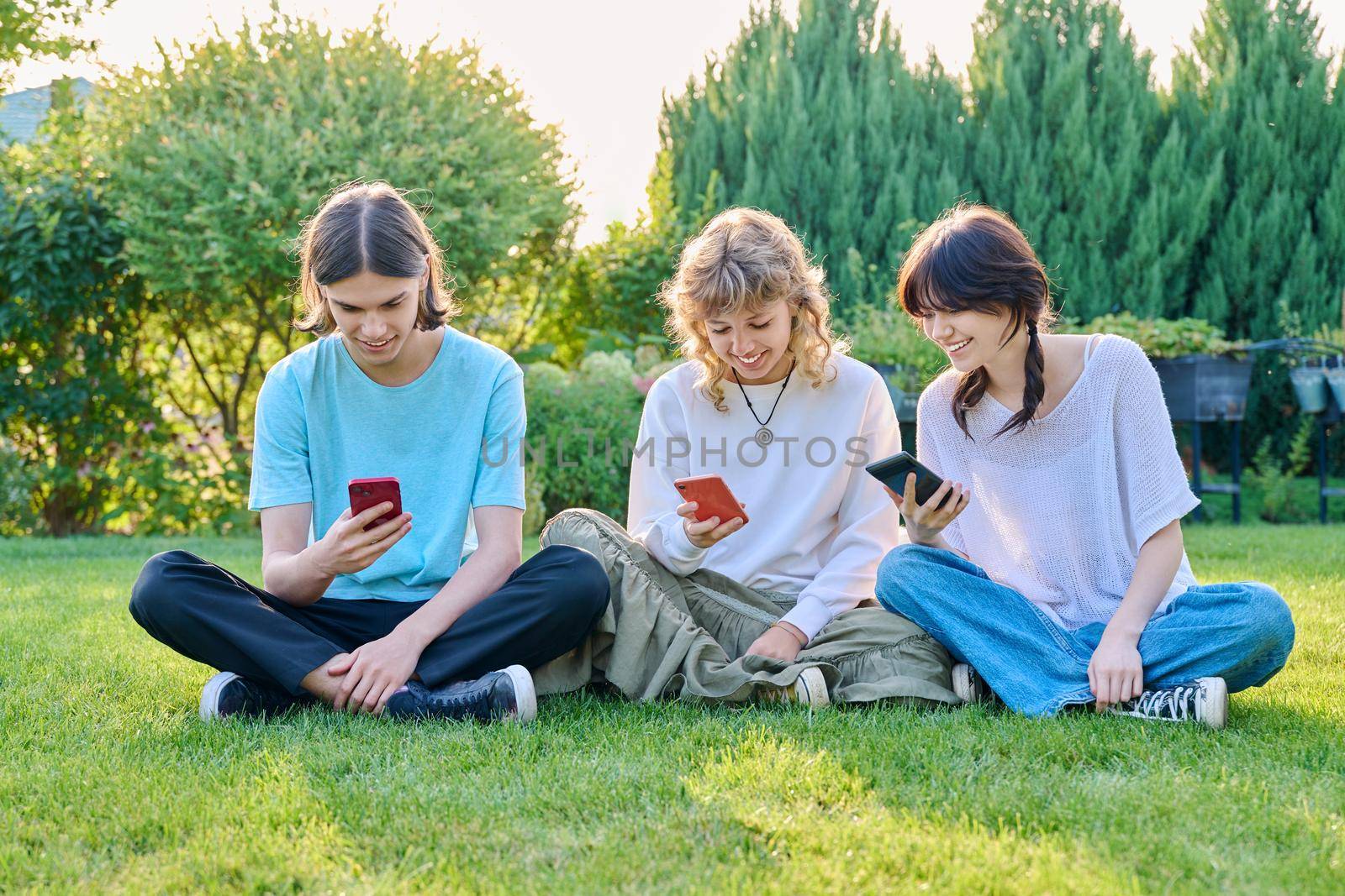 Group of teenagers sitting on grass with smartphones. Three teenage friends relaxing on lawn, sunny summer day, using mobile applications for leisure. Lifestyle technology and friendship youth concept
