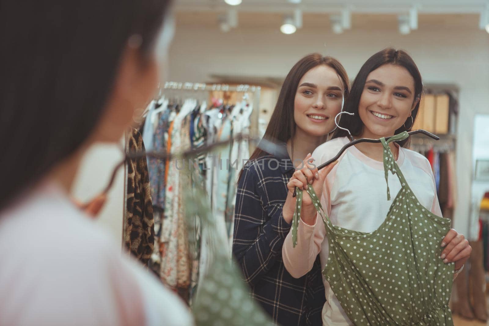 Cheerful beautiful woman smiling, helping her best friend choosing a new dress to buy. Two female friends examining a dress in front of the mirror at clothing store. Gorgeous woman shopping with friend
