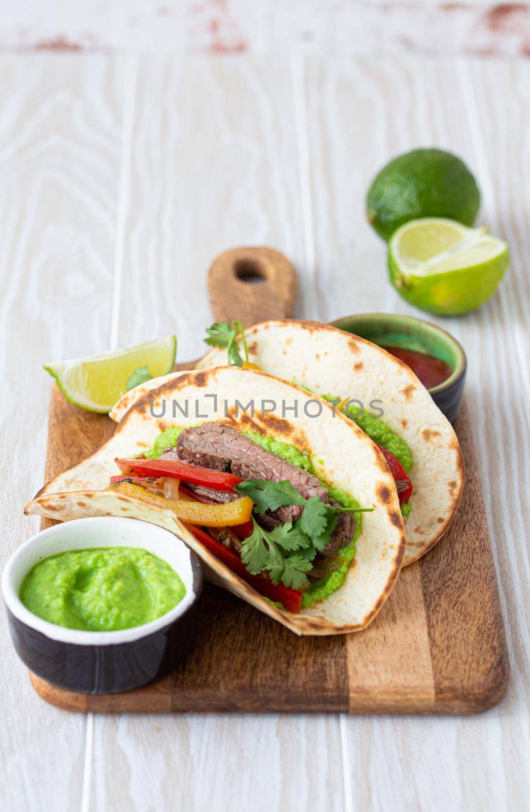Traditional Mexican dish Beef fajitas tacos served on wooden cutting board with tomato salsa and guacamole on rustic white wooden background from angle view, American Mexican food