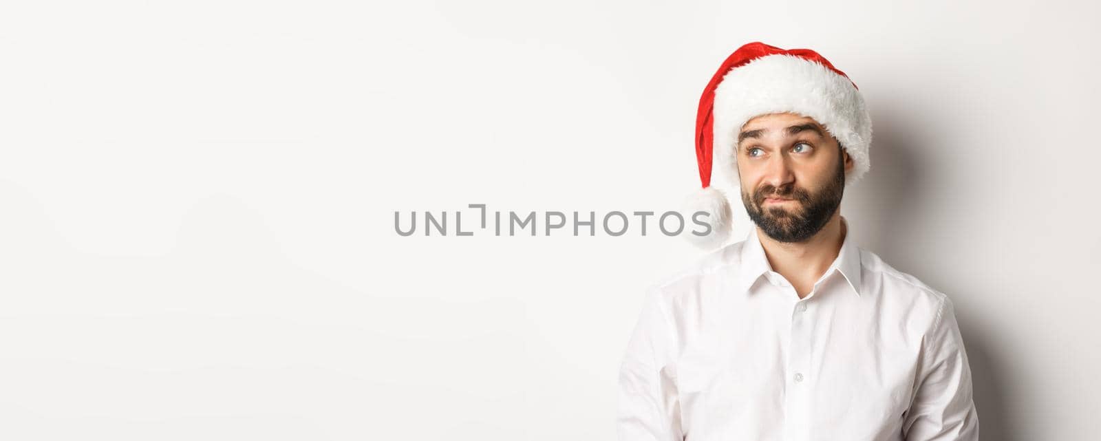 Close-up of skeptical guy in santa hat looking doubtful left, grimacing with hesitation, white background.