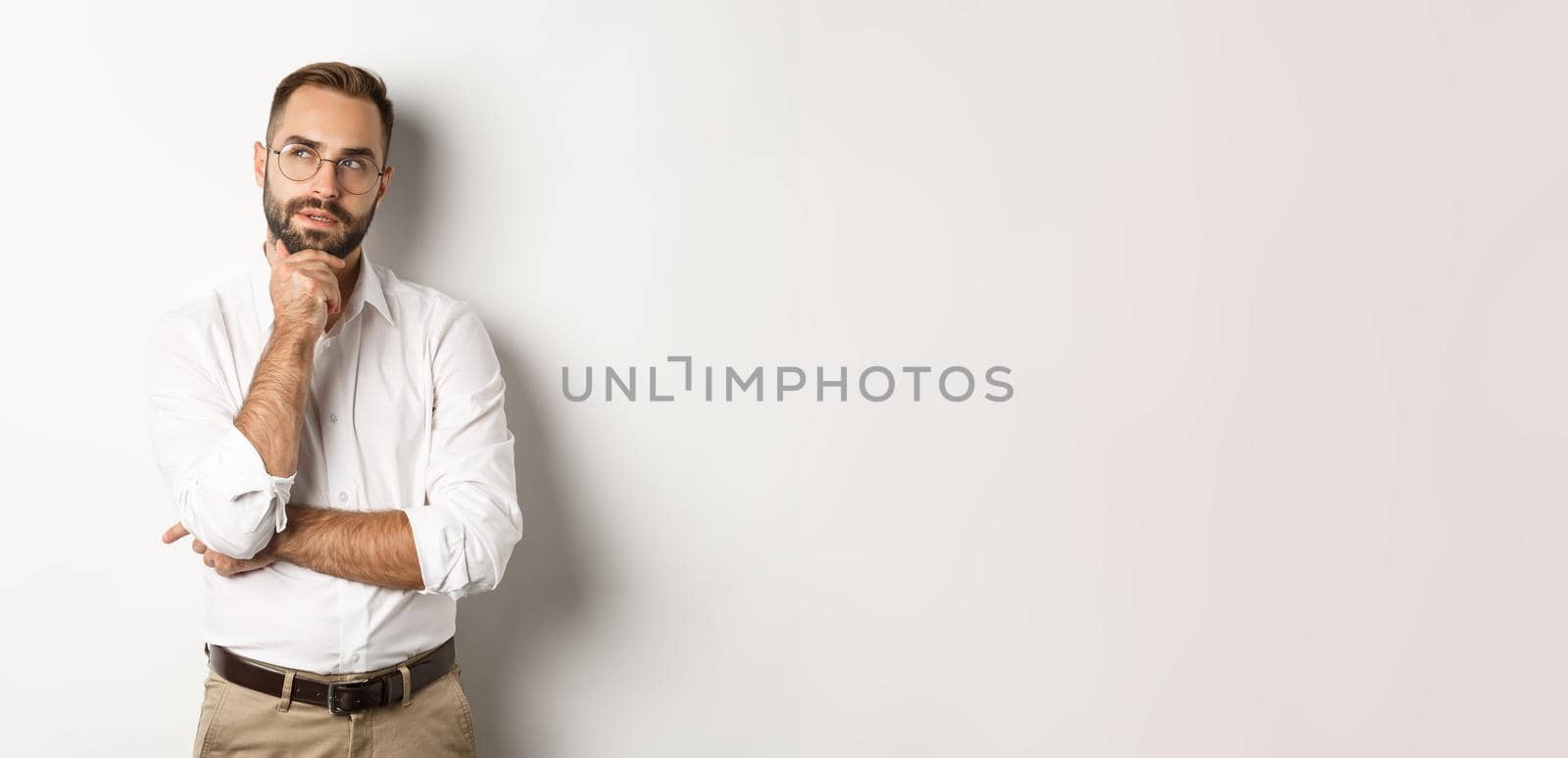 Thoughtful businessman in glasses making plan, looking at upper left corner and thinking, standing against white background.