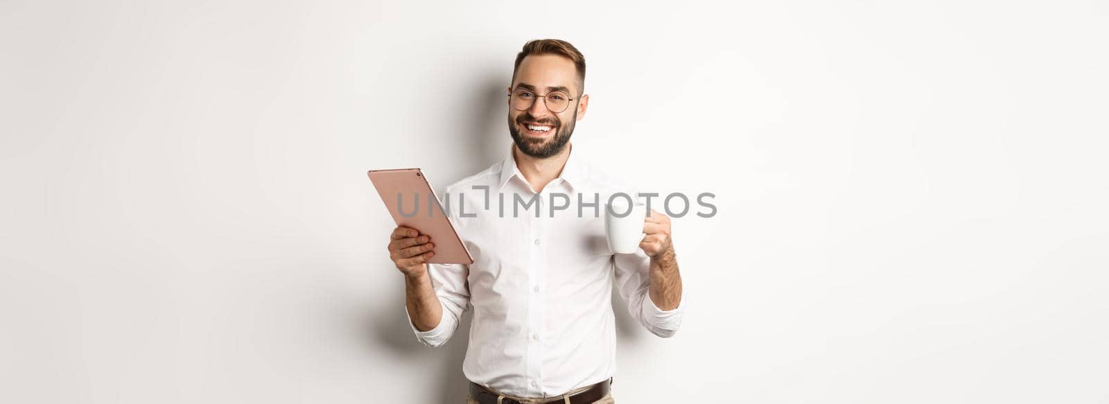 Satisfied boss drinking tea and using digital tablet, reading or working, standing over white background.