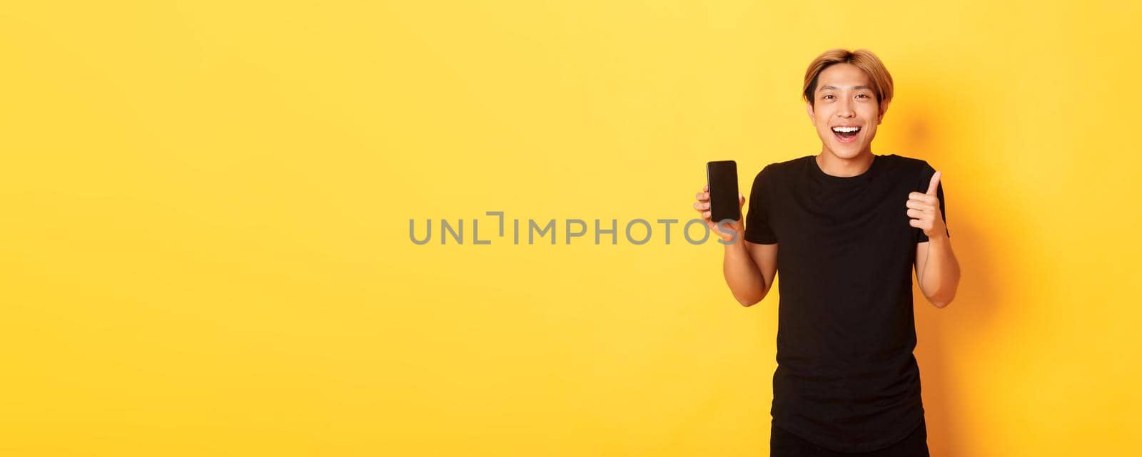 Portrait of pleased smiling asian guy, showing smartphone screen and thumbs-up satisfied with application, standing yellow background by Benzoix