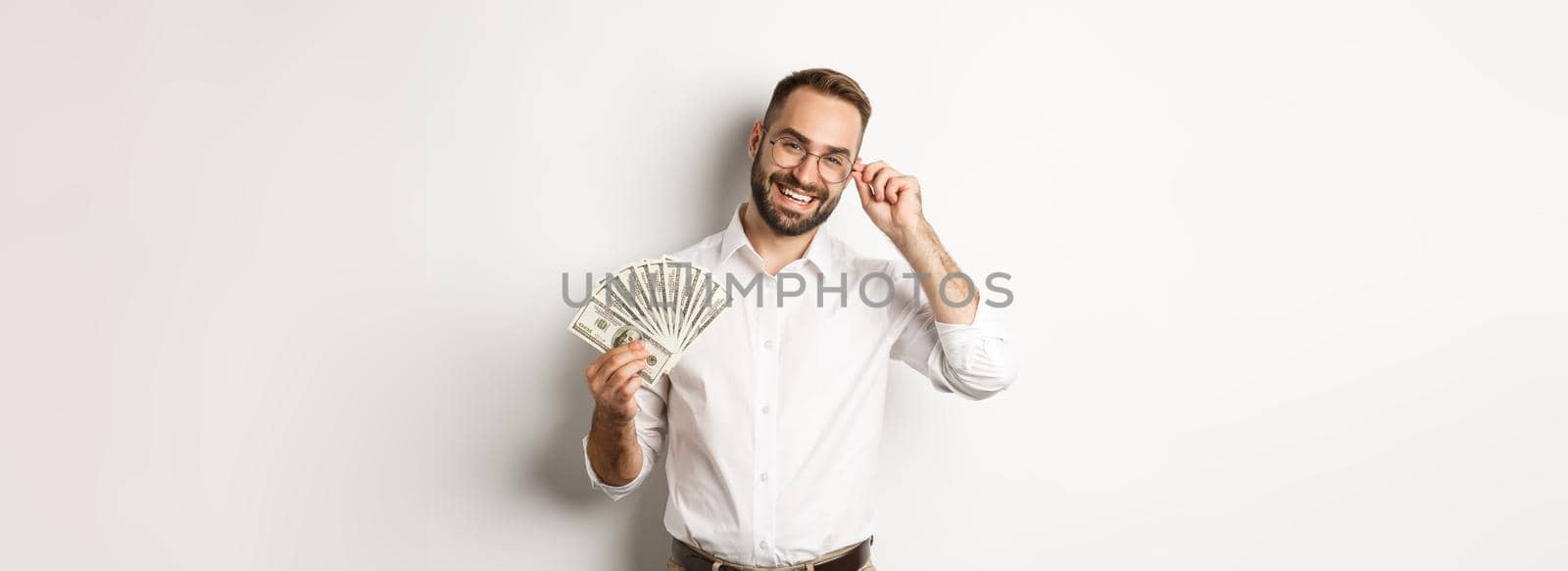 Handsome successful businessman holding money, fixing glasses on nose, standing over white background.