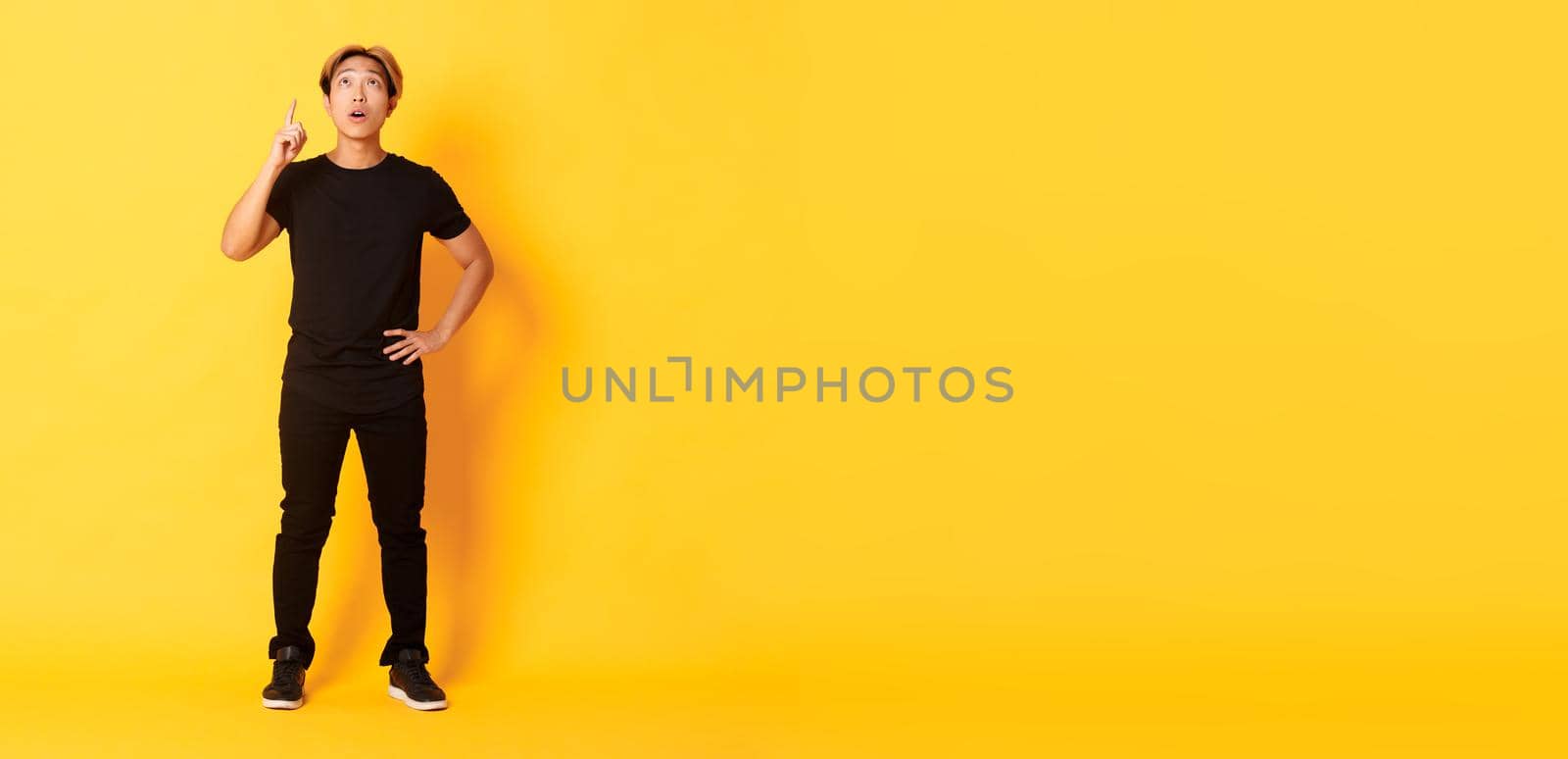 Full-length of thoughtful, intrigued attractive blond asian man raising finger, pointing up and looking amused, yellow background.