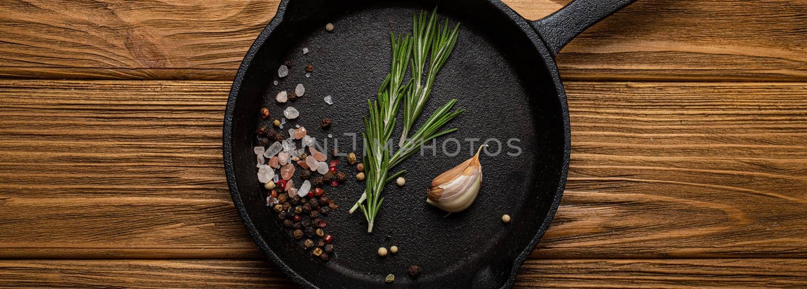 Black cast iron frying pan skillet with food cooking ingredients fresh rosemary, garlic, salt and pepper on rustic wooden table, cooking background and healthy eating kitchen concept