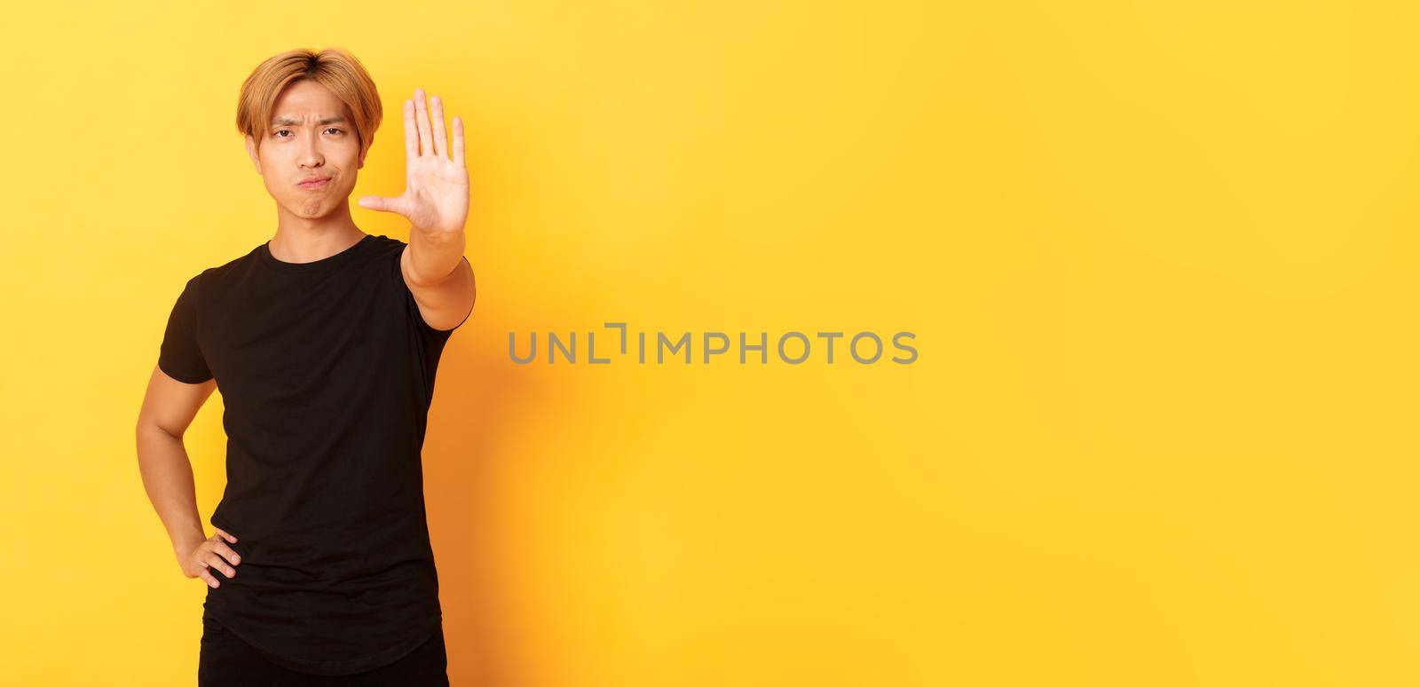 Portrait of disappointed serious asian man, smirking displeased and extend hand, showing stop gesture, yellow background by Benzoix