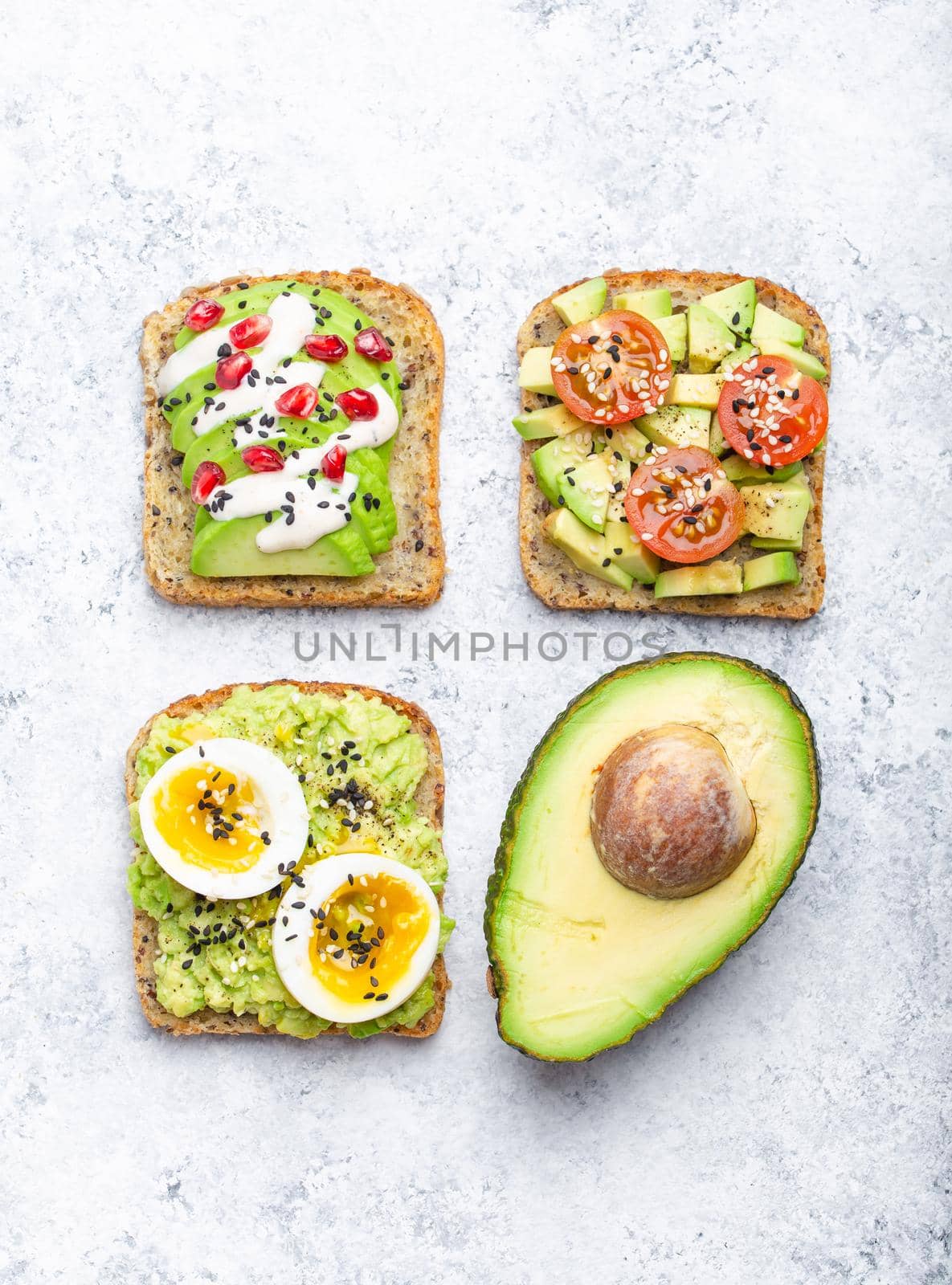 Avocado toasts with egg, tomatoes, seasonings and a half of whole avocado over white stone background. Healthy breakfast avocado sandwiches with different toppings, top view, close-up.