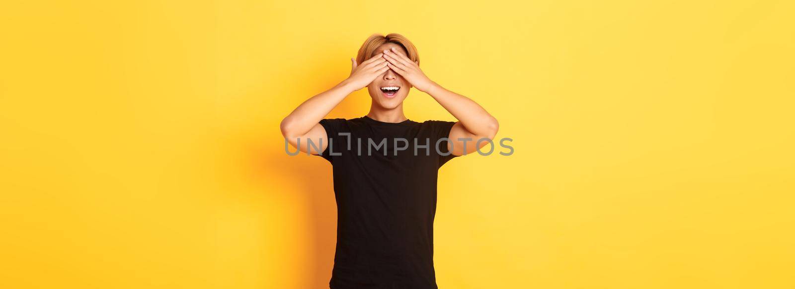 Portrait of amused and excited smiling blond guy shut eyes with palms, waiting for surprise, standing yellow background by Benzoix