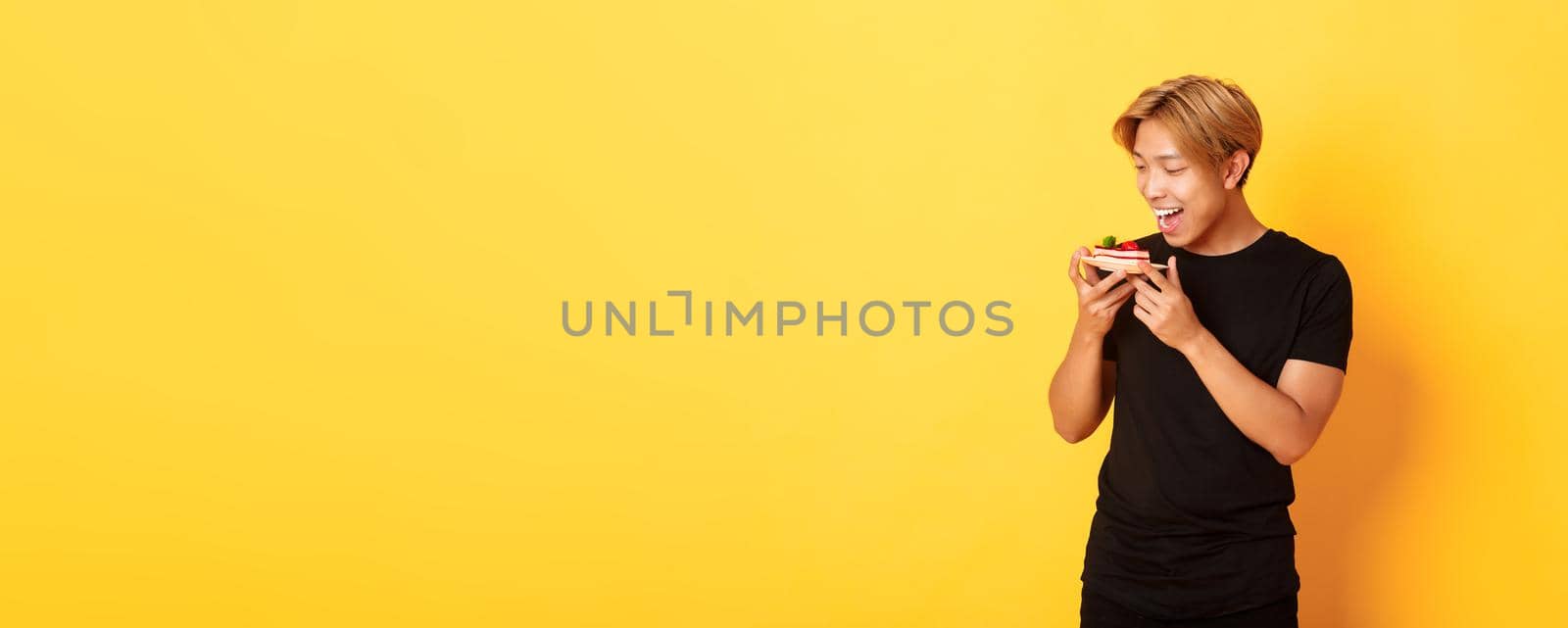 Joyful handsome asian guy smiling and looking tempted at cake while making bite, standing yellow background.