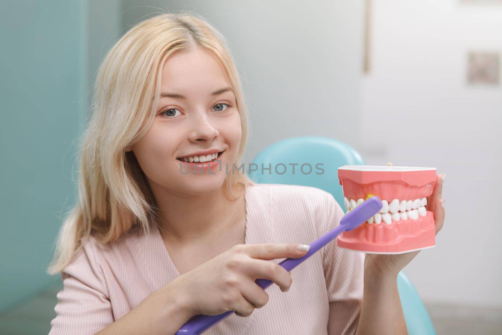 Cheerful young woman smiling to the camera while showing how to brush teeth correctly on a jaw model