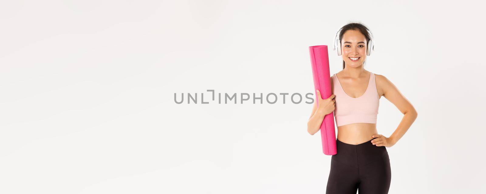 Sport, wellbeing and active lifestyle concept. Sassy beautiful asian fitness girl, woman ready for yoga classes or home training, holding rubber mat for workout and listening music in headphones.