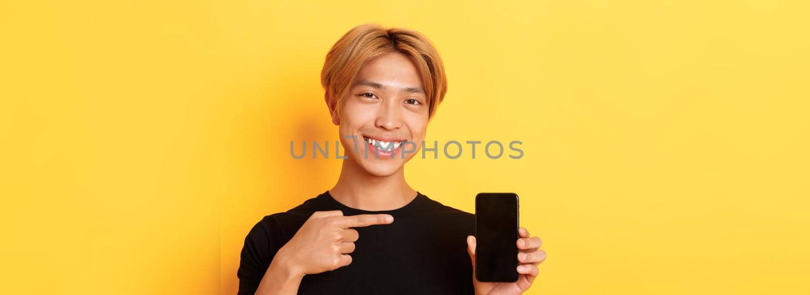 Cheerful attractive korean guy pointing finger at mobile phone screen, smiling pleased, standing over yellow background.