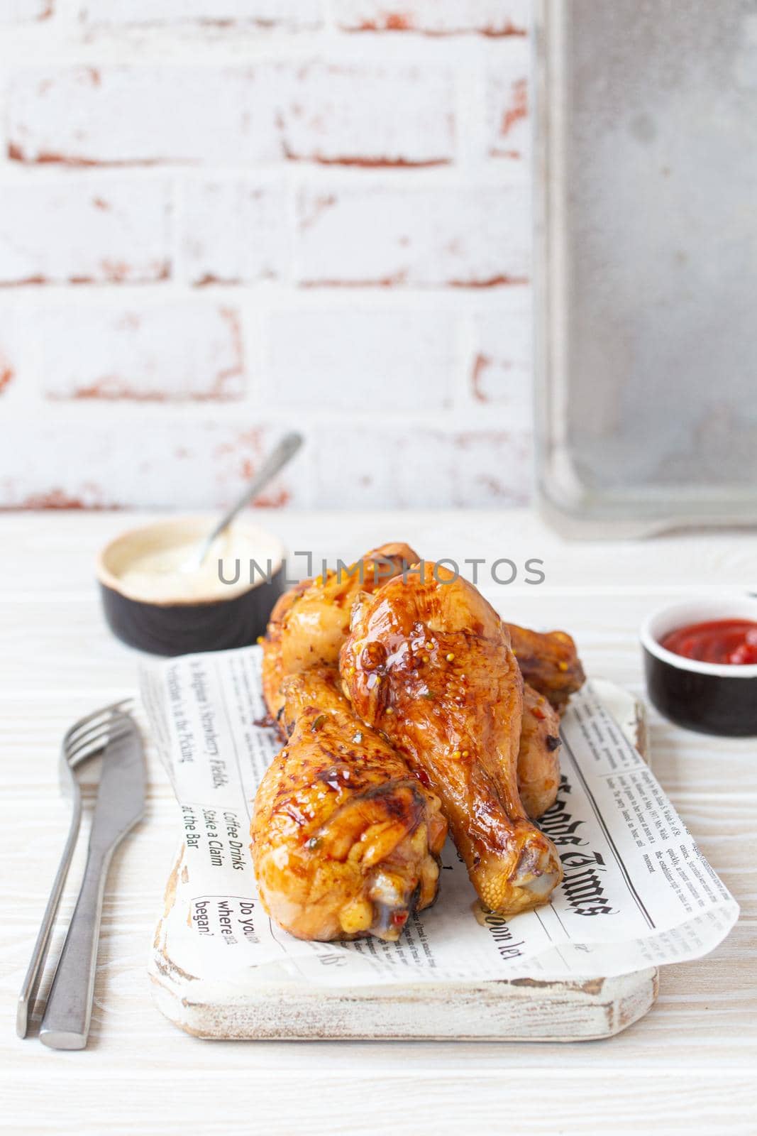 Roasted chicken legs drumsticks on wooden board with newspaper sheet served with ketchup and mayonnaise on rustic white wooden table, angle view