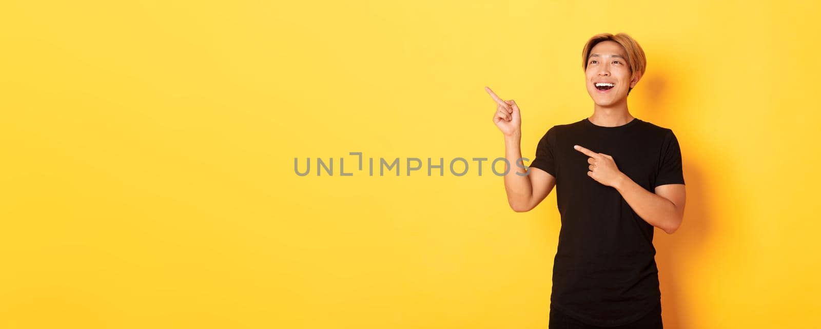 Portrait of pleased handsome asian guy in black t-shirt, pointing fingers and looking upper left corner with satisfied smile, yellow background by Benzoix