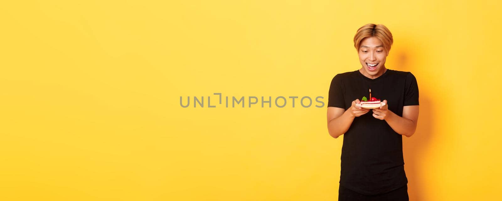 Excited happy attractive asian guy smiling as looking at birthday cake, making wish, celebrating b-day, standing yellow background.