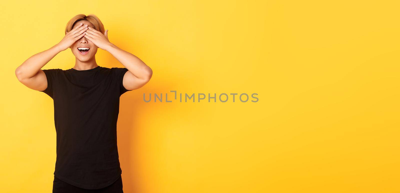 Portrait of amused and excited smiling blond guy shut eyes with palms, waiting for surprise, standing yellow background.