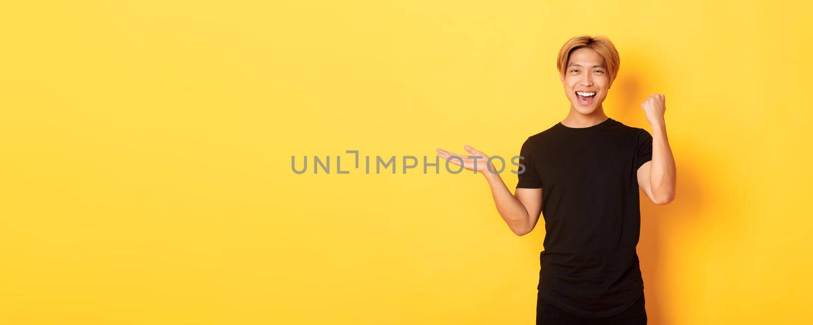 Satisfied and happy attractive korean guy smiling, fist pump with rejoice, holding something on hand, standing yellow background.
